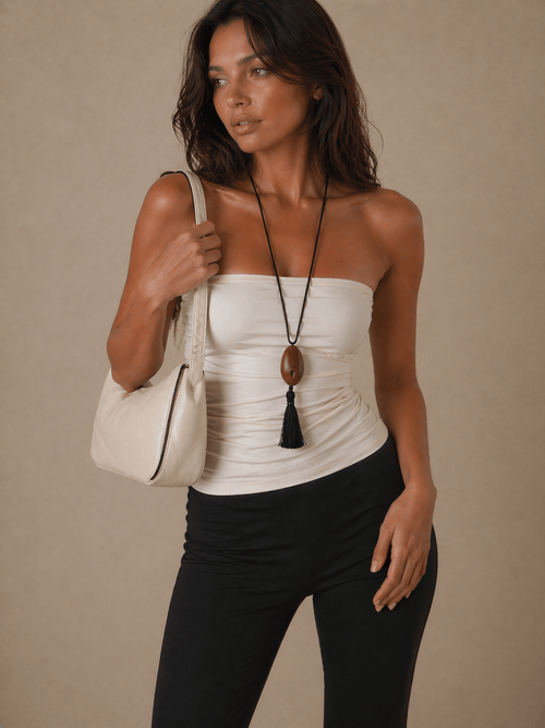 Woman wearing a white strapless top, a necklace with a wooden pendant and black tassel and black pants, holding a beige handbag against a beige background.