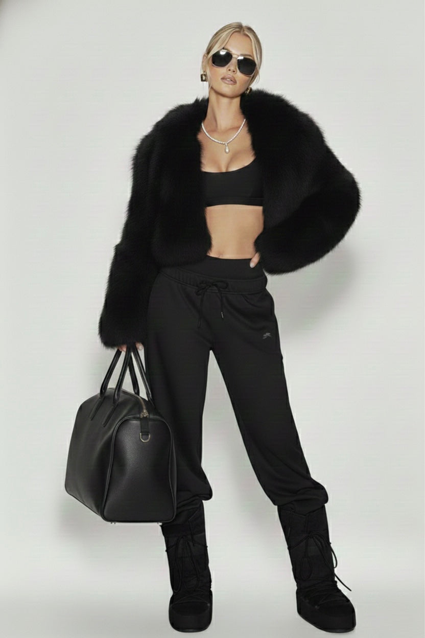 A woman wearing a black fur coat, a tennis necklace with a teardrop pearl pendant, Josephine Necklace by Vintaras, black sports bra, black pants, and black boots, holding a black bag on a white background.
