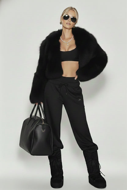 A woman wearing a black fur coat, a tennis necklace with a teardrop pearl pendant, Josephine Necklace by Vintaras, black sports bra, black pants, and black boots, holding a black bag on a white background.