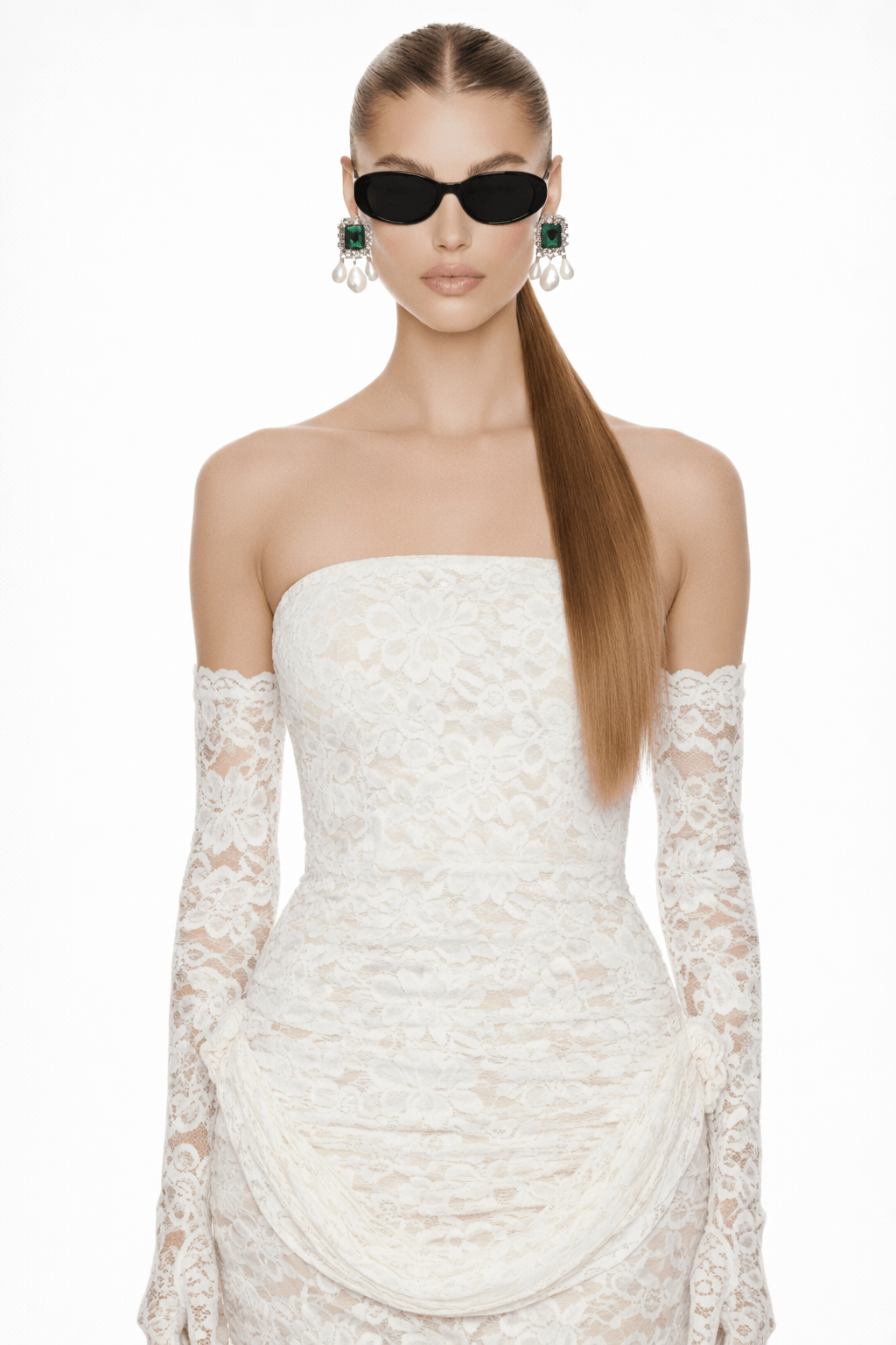 A bride wearing green gemstone earrings with pearl drops, a white bridal dress and lace gloves, and holding  a shell-shaped clutch with a plain background.