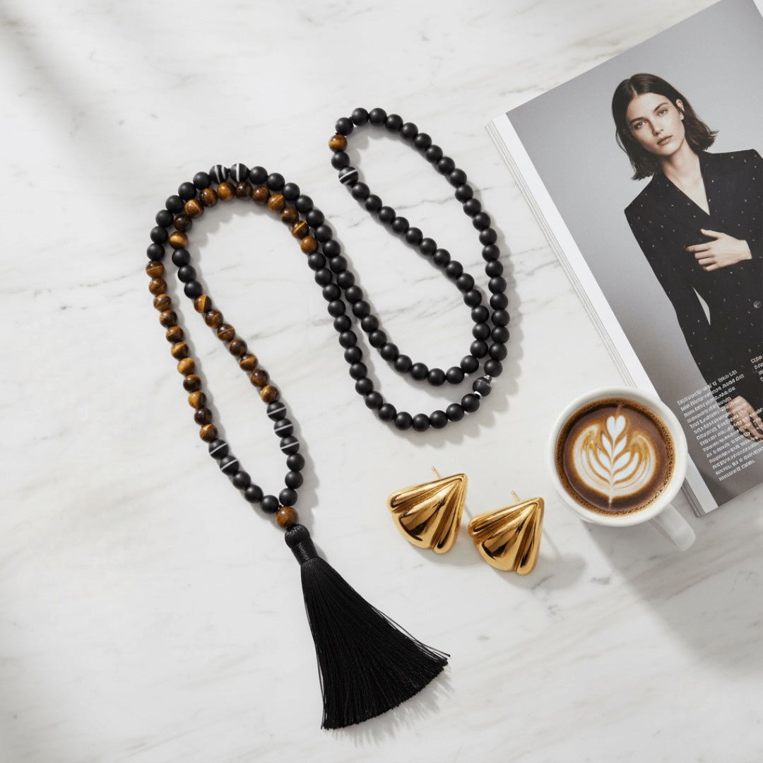 Black tassel necklace with tiger eye beads , gold triangle earrings, and a coffee cup on a magazine with a woman's face.