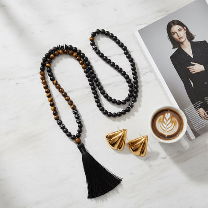 Black tassel necklace with tiger eye beads , gold triangle earrings, and a coffee cup on a magazine with a woman's face.