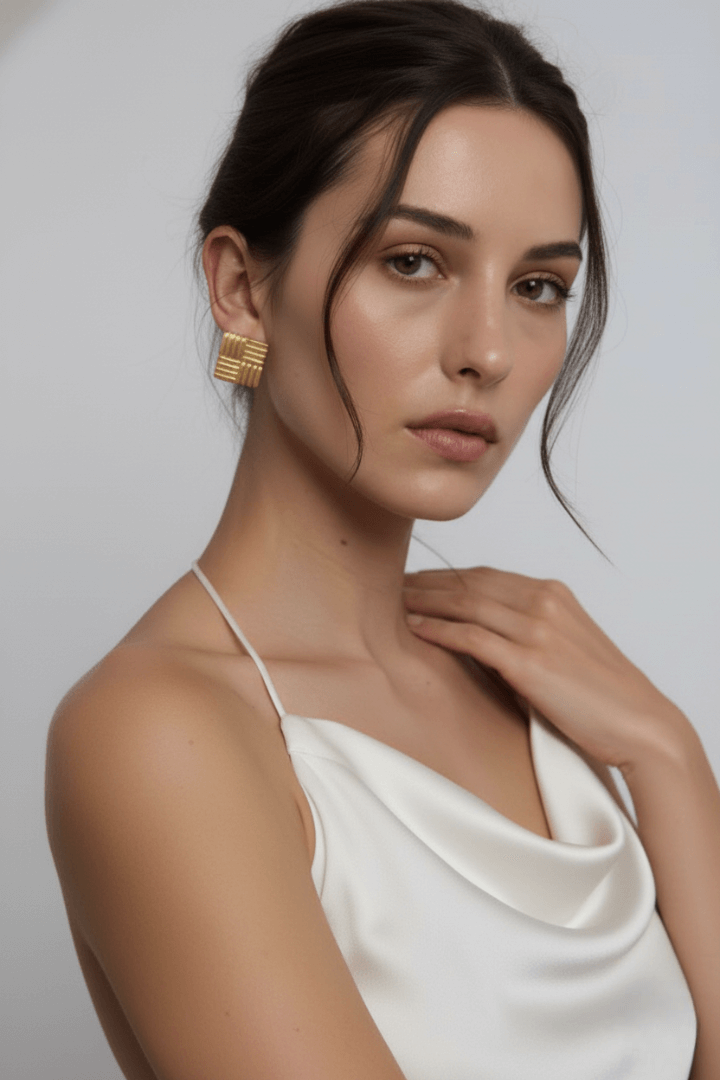Woman wearing geometric gold square earrings from Vintaras jewelry and a white top against a plain background