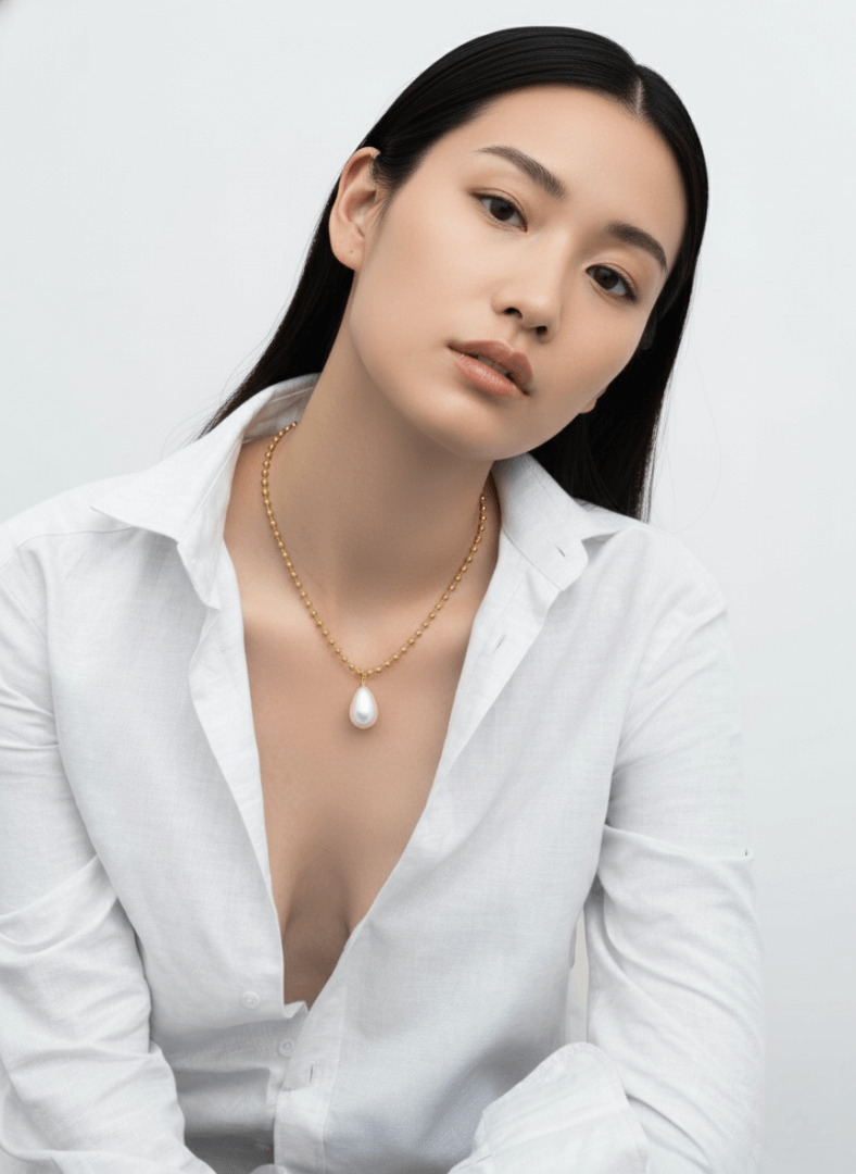 Woman wearing a white blouse and gold Vintaras Ingrid Necklace with tear drop pearl pendant against a plain background