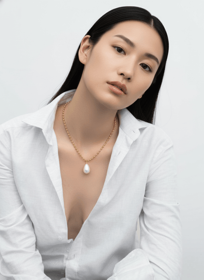 Woman wearing a white blouse and gold Vintaras Ingrid Necklace with tear drop pearl pendant against a plain background